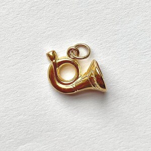 May include: A gold-colored charm in the shape of a French horn. The charm has a loop at the top for attaching to a necklace or bracelet. The horn is detailed with a bell and a mouthpiece. The charm is likely made of metal and has a shiny finish.