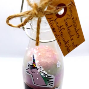 Könnte beinhalten: Eine kleine Glasflasche gefüllt mit rosa und weißen Badesalzen, verziert mit einem Einhorn-Aufkleber und einem Etikett mit der Aufschrift "Handgemachte Badesalze mit Mineralölen".