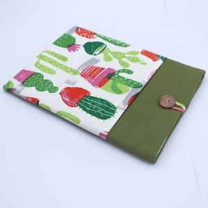 Puede incluir: Una funda para portátil de tela blanca y verde con estampado de cactus. La funda tiene una solapa verde con cierre de botón marrón.