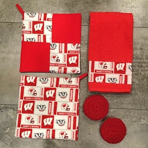 May include: Set of three red and white kitchen towels with the University of Wisconsin Badgers logo. The set includes two pot holders and one hand towel. The pot holders are square and the hand towel is rectangular. The towels are made of a soft, absorbent fabric. The set also includes two red crocheted scrubbers.
