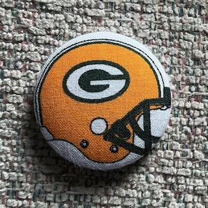 May include: A round button featuring a football helmet design in green, white, and orange. The helmet has a green "G" logo on the side. The button is made of fabric and has a white border.