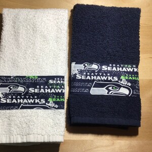 May include: Two white bath towels with a green and white Seattle Seahawks logo printed on the bottom edge. The logo includes the words "Seattle Seahawks" and a stylized image of a hawk.