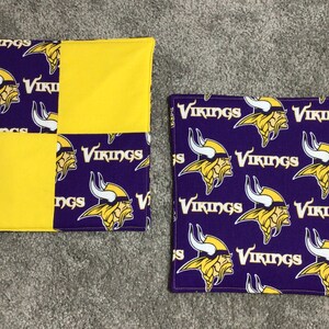 May include: Two purple and yellow pot holders with the Minnesota Vikings logo. The pot holders are made of fabric and have a loop for hanging.