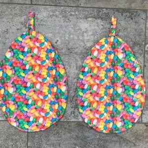 May include: Two colorful pot holders shaped like eggs with a jelly bean pattern. The pot holders are made of fabric and have a loop for hanging.