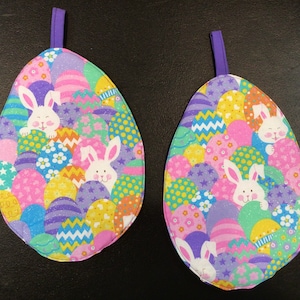 May include: Two purple and white egg-shaped pot holders with a colorful Easter egg and bunny rabbit design.
