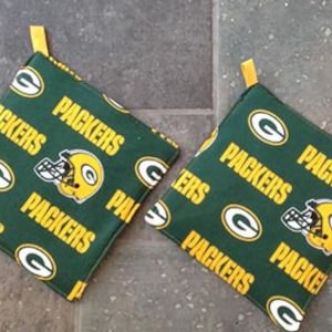 May include: Two green and yellow fabric pot holders with the word "Packers" repeated on them. The pot holders have a green and yellow football helmet graphic.