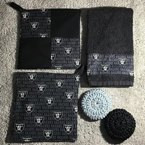 May include: A set of black and silver Raiders themed bathroom accessories. The set includes a hand towel, two pot scrubbers, and two pot holders. The pot holders and hand towel feature a repeating pattern of the Raiders logo and the word "RAIDERS".