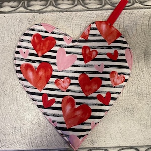 May include: A black and white striped heart-shaped pot holder with red and pink hearts printed on it. The pot holder has a loop at the top for hanging.
