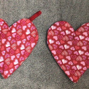 May include: Two heart-shaped oven mitts. The mitts are red with a pattern of pink and white hearts. Each mitt has a red loop for hanging.