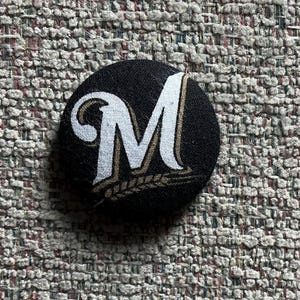 May include: A black button with a white and gold letter "M" design. The button has a fabric texture and a circular shape. The letter "M" is stylized with a decorative serif font and a wheat-like design.