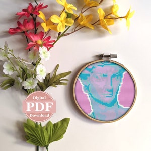 Puede incluir: Un patrón de punto de cruz de un busto azul y verde sobre un fondo rosa en un aro de bordado de madera. El patrón está etiquetado como "Descarga digital PDF".