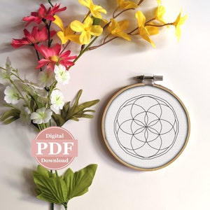 May include: A black and white geometric pattern in a circular embroidery hoop. The pattern is a flower of life design. The hoop is on a white background with flowers.