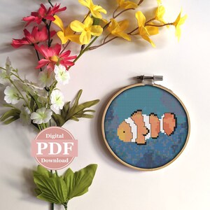 May include: A cross-stitch pattern of an orange and white clownfish swimming in blue water. The pattern is in a wooden embroidery hoop.