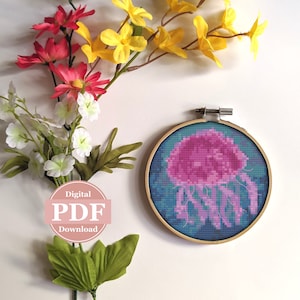 May include: A digital PDF download pattern for cross-stitching a pink jellyfish with blue and green background. The pattern is displayed in a wooden embroidery hoop.