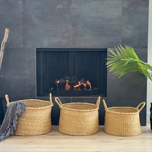 May include: Three large woven baskets with handles in front of a fireplace. The baskets are made of natural materials and have a light brown color. The fireplace is black and has a fire burning inside.