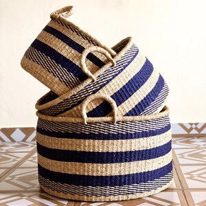 May include: Three woven baskets with blue and natural colored stripes. The baskets are stacked on top of each other. The largest basket is on the bottom and the smallest basket is on top.