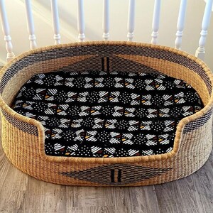 May include: A woven wicker pet bed with a black and white patterned cushion. The bed is oval shaped and has a raised rim.