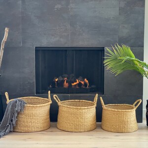 May include: Three large woven baskets with handles in front of a fireplace. The baskets are made of natural materials and have a light brown color. The fireplace is black and has a fire burning inside.