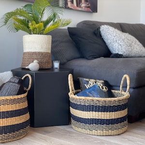 May include: Two large woven baskets with black and natural colored stripes and handles. The baskets are sitting on a wooden floor in front of a gray couch. One basket is filled with a book titled "The Times Atlas of the World".