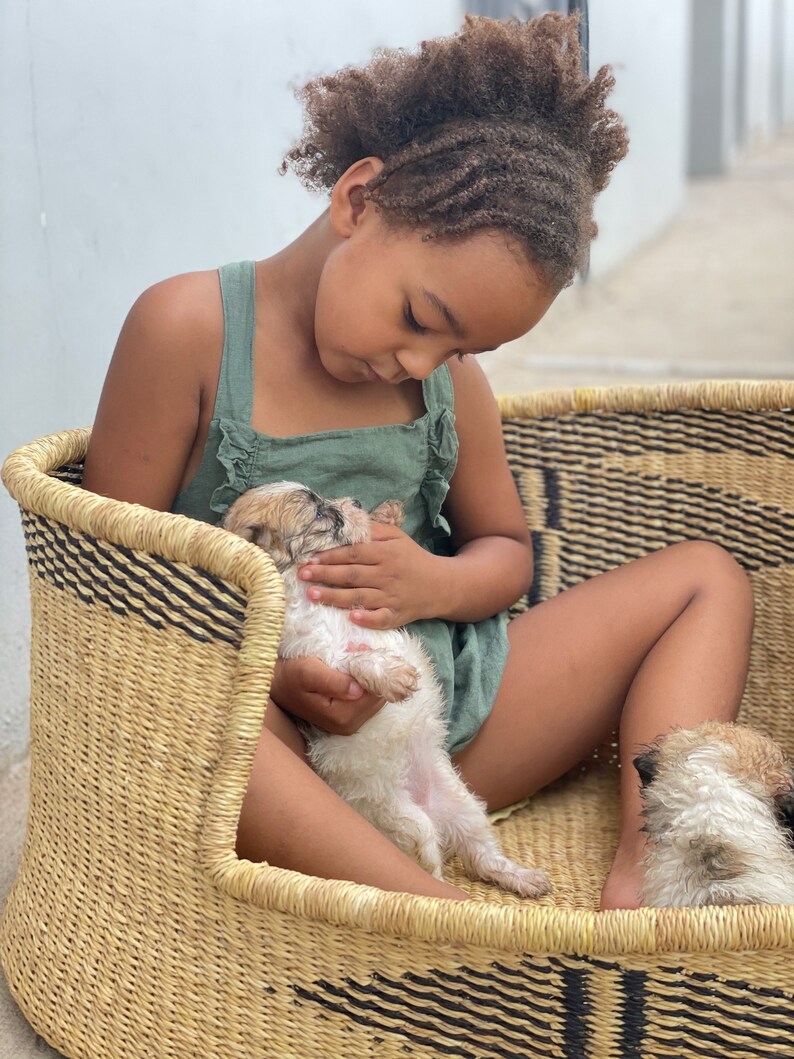 May include: A young girl with curly brown hair sits in a woven basket holding a small white puppy. The puppy is lying on its back with its paws in the air. The girl is wearing a green sleeveless dress.
