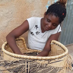 May include: A woman is weaving a large, round, natural fiber basket with black and white stripes. The basket is almost finished and has a fringe around the top edge.