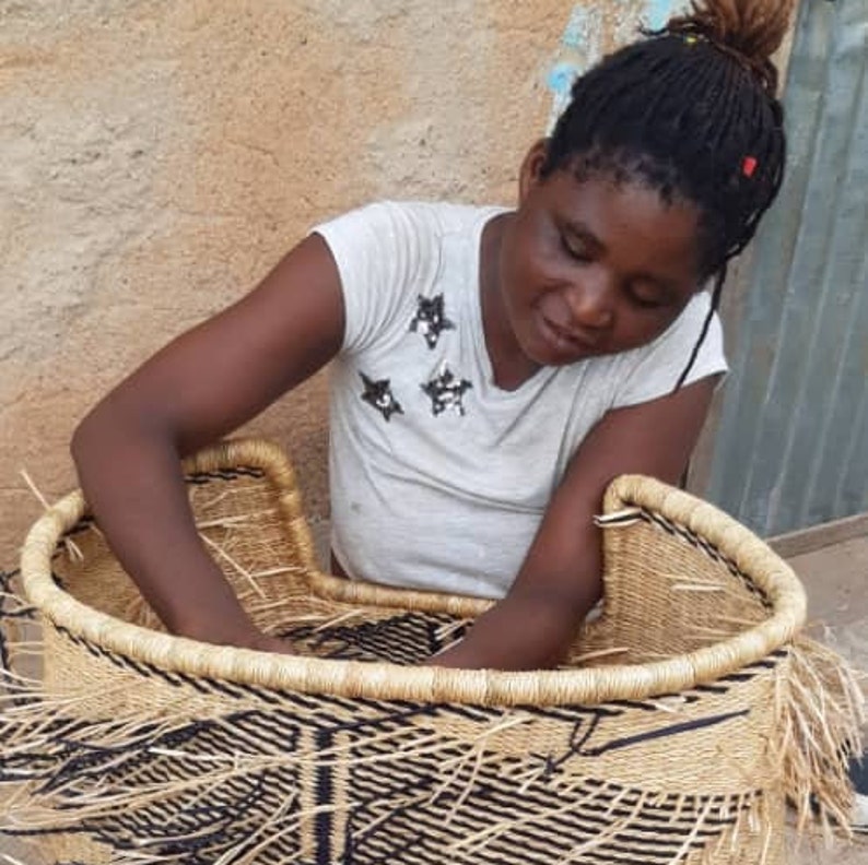 May include: A woman is weaving a large, round, natural fiber basket with black and white stripes. The basket is almost finished and has a fringe around the top edge.