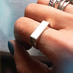 May include: A silver rectangular ring worn on a finger with several other silver rings.