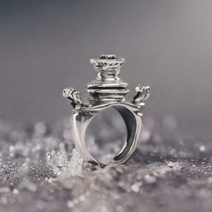 May include: A silver ring with a detailed, ornate design. The ring features a central, tiered structure with decorative elements, possibly inspired by a historical or architectural theme. The ring is set against a blurred, textured background.