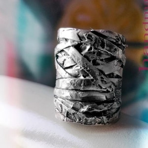 May include: A silver ring with a textured, layered design. The ring is handmade and has a unique, organic look.