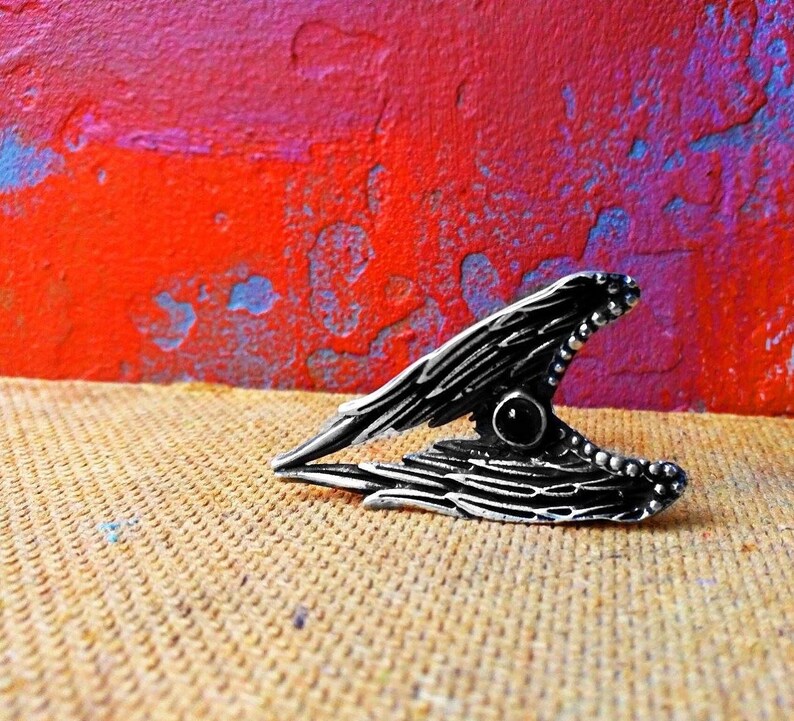 May include: A silver ring featuring a black gemstone at its centre, shaped like a wing with intricate detailing.
