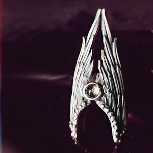 May include: A silver ring featuring a clear gemstone set in the centre, shaped like two wings meeting.