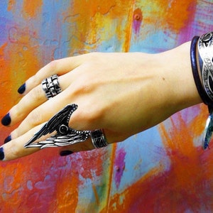 May include: A hand wearing a silver ring with a wing design and a silver band ring. The hand is also wearing a purple leather bracelet with silver accents.