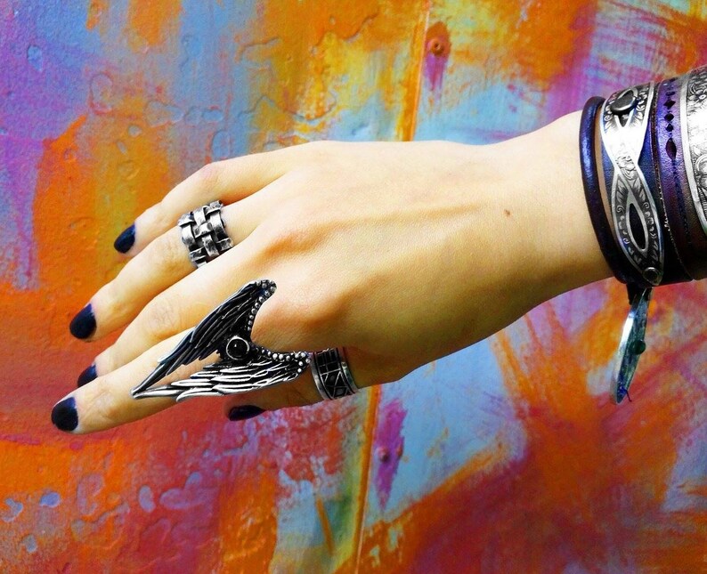 May include: A hand wearing a silver ring with a wing design and a silver band ring. The hand is also wearing a purple leather bracelet with silver accents.