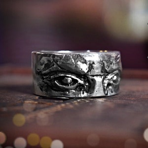 May include: A silver ring with a detailed design of two eyes. The ring is wide and has a textured surface.