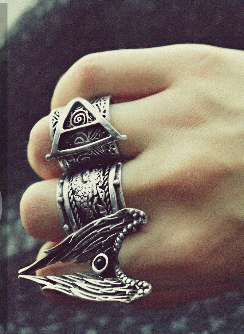 May include: Three silver rings worn on a finger. The top ring features a triangle with a spiral design. The middle ring has a braided design. The bottom ring is a winged design with a black stone in the centre.