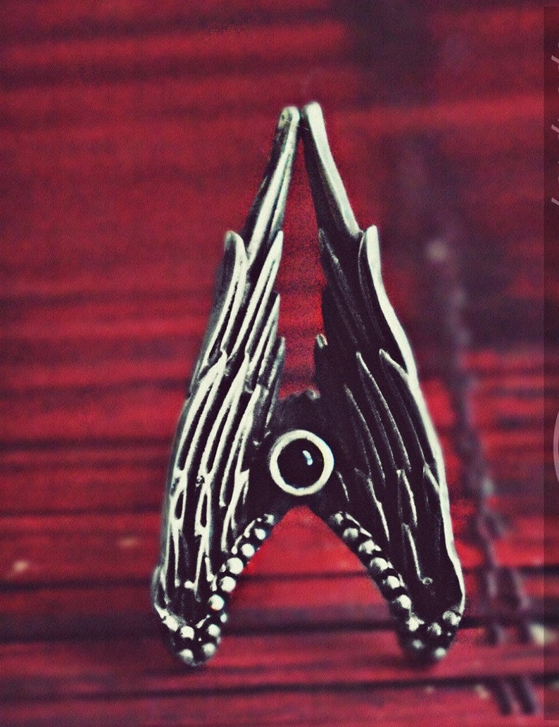 May include: A silver ring with a black stone in the centre and two wings on either side. The ring is on a red background.