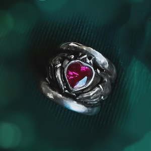 Puede incluir: Un anillo plateado con una piedra preciosa en forma de corazón de color rubí. El anillo tiene un diseño detallado y texturizado alrededor de la piedra preciosa, sobre un fondo verde oscuro con luces bokeh borrosas.