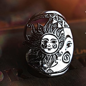 Può includere: Anello color argento con un design sole e luna. Il sole ha una faccia sorridente con raggi, e la luna ha una faccia con un occhio e una spirale. L'anello è dettagliato con motivi intricati.