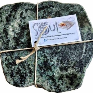 May include: A dark green granite charcuterie board with a natural, irregular shape. The board is tied with twine and includes a small white tag with the text "STONE SOUL" and contact information. The granite has a speckled pattern.