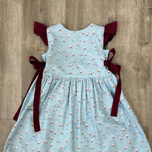 May include: A light blue dress with a white and red print of small figures playing musical instruments. The dress has maroon ribbon ties at the sides and maroon ruffled sleeves.