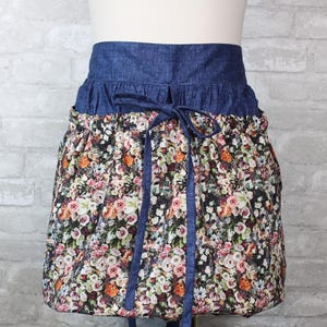 May include: A waist apron featuring a dark blue denim waistband with a matching tie closure. The lower portion of the apron is made of a gathered fabric with a dense, multi-colored floral print consisting of pink, orange, white, and green flowers.