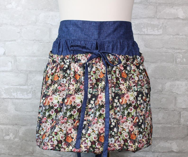 May include: A waist apron featuring a dark blue denim waistband with a matching tie closure. The lower portion of the apron is made of a gathered fabric with a dense, multi-colored floral print consisting of pink, orange, white, and green flowers.