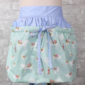 May include: A gathered apron featuring a light blue waistband and a mint green skirt with a repeating pattern of white farm animals, including cows, pigs, and chickens, adorned with colorful floral accents and white windmill illustrations.