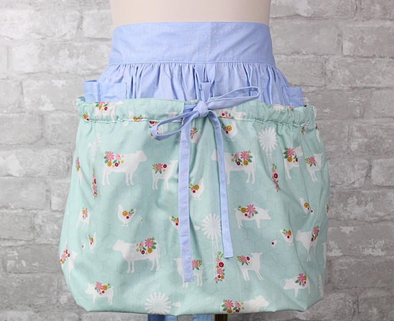 May include: A gathered apron featuring a light blue waistband and a mint green skirt with a repeating pattern of white farm animals, including cows, pigs, and chickens, adorned with colorful floral accents and white windmill illustrations.