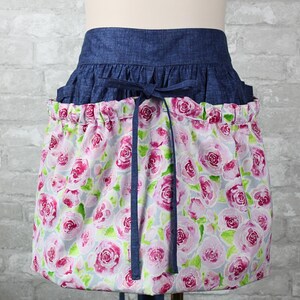 May include: A waist apron featuring a dark blue denim waistband with a tie closure and a gathered skirt section. The skirt is made of a light gray fabric printed with a repeating pattern of pink and magenta roses with green leaves.