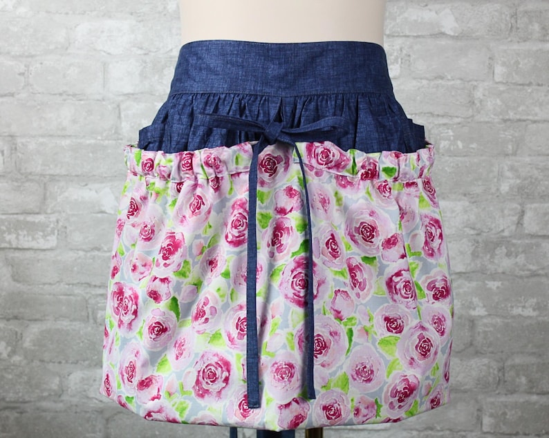 May include: A waist apron featuring a dark blue denim waistband with a tie closure and a gathered skirt section. The skirt is made of a light gray fabric printed with a repeating pattern of pink and magenta roses with green leaves.