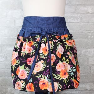 May include: A half apron with a denim blue waistband and ties. The lower portion of the apron features a black background with a floral pattern of orange, pink, and green flowers. The apron is gathered at the waist.