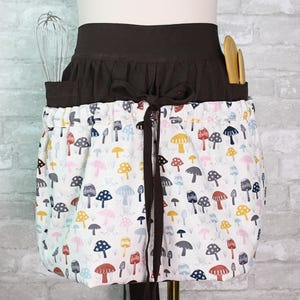 May include: A waist apron featuring a dark brown waistband with a tie closure and a white skirt section patterned with colorful mushroom illustrations. The apron includes two pockets containing a metal whisk and two wooden kitchen utensils.