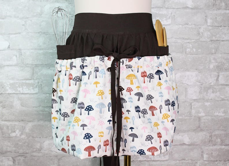 May include: A waist apron featuring a dark brown waistband with a tie closure and a white skirt section patterned with colorful mushroom illustrations. The apron includes two pockets containing a metal whisk and two wooden kitchen utensils.