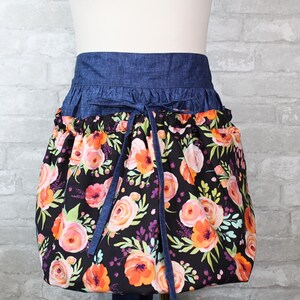May include: A waist apron featuring a dark blue denim waistband with matching tie strings and a gathered skirt section. The skirt is made of black fabric printed with a vibrant floral pattern in shades of orange, pink, and light green foliage.